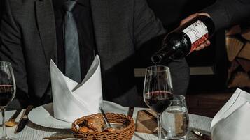 Eine Person gießt Wein in ein Glas an einem Esstisch, auf dem ein gebratenes Entenoberstück, Brot und andere Speisen stehen.