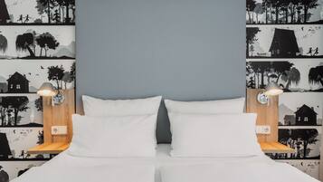 A double bed with white linens, two bedside lamps, and a wall with a nature-themed wallpaper.