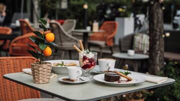 Un área de comedor al aire libre con sillas de mimbre, una mesa con platos de comida, una planta en maceta y un vaso de postre.
