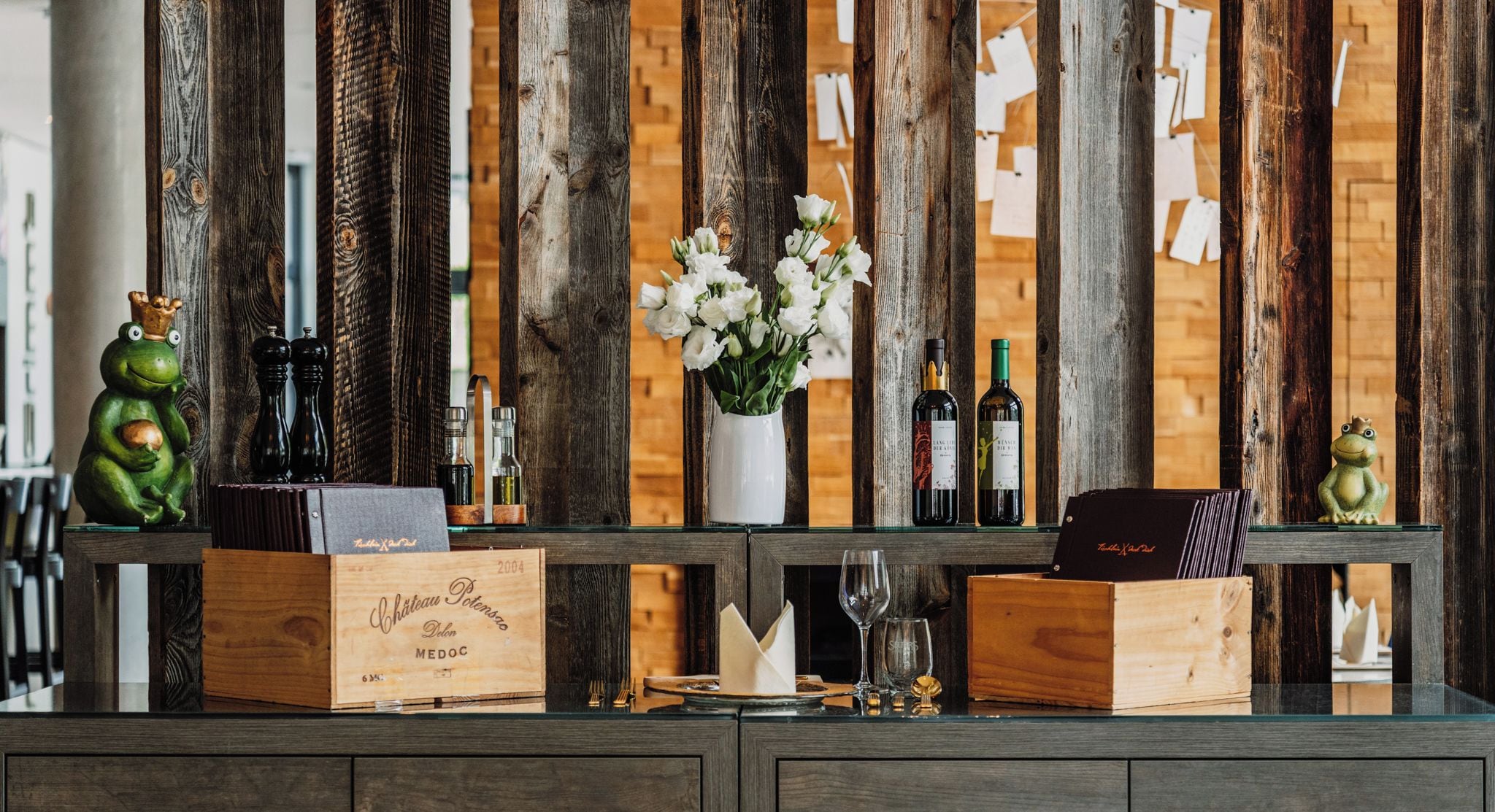 A wooden display with wine bottles, a vase of flowers, and decorative frog figurines.