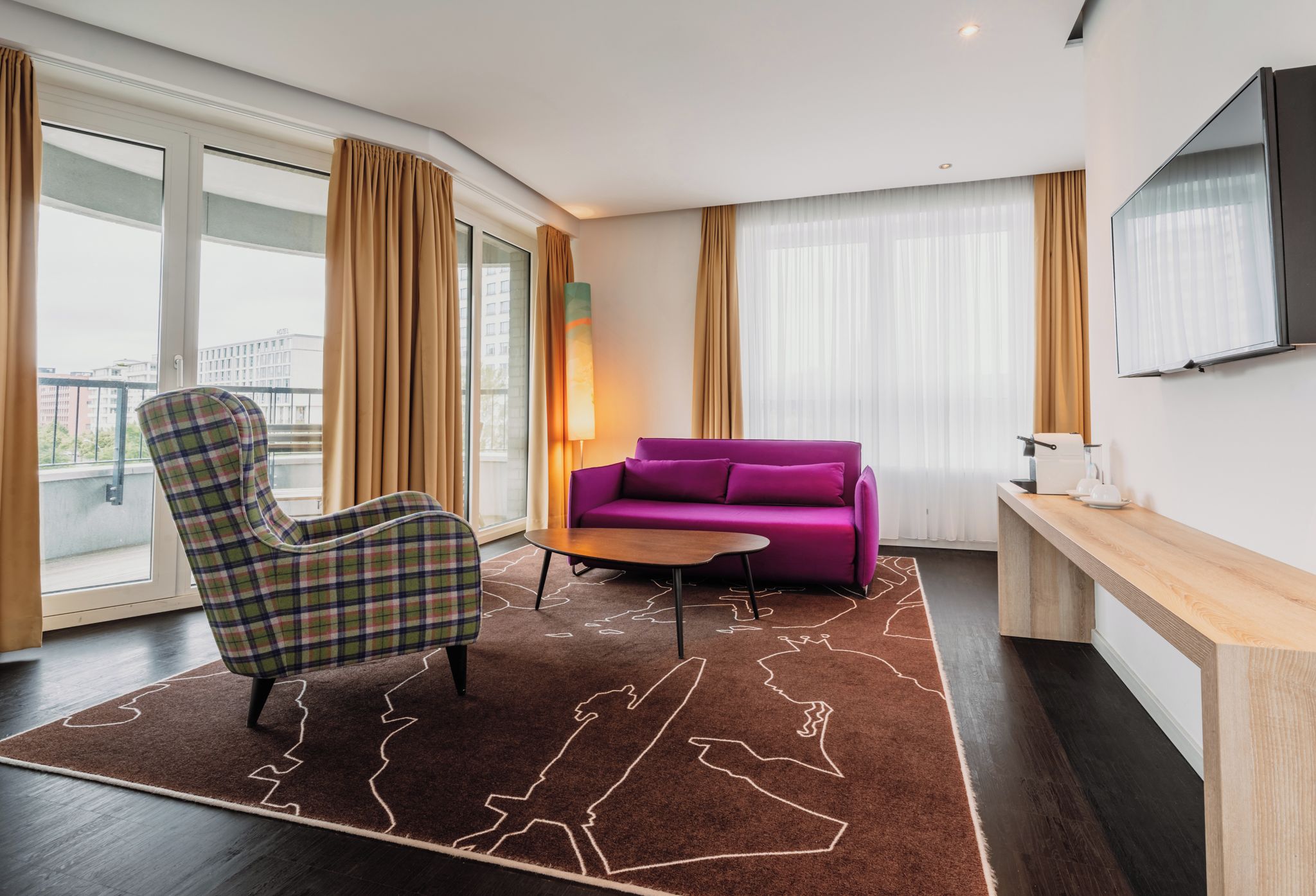 Hotellihuone, jossa on suuri ikkuna, sohva, tuoli, pieni pöytä ja seinälle asennettu televisio.