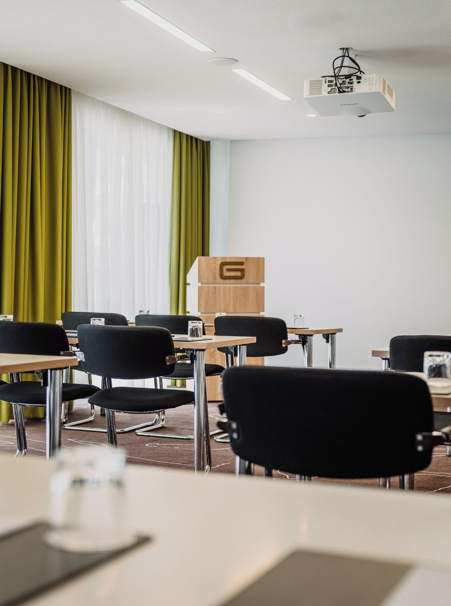 A conference room with a projector, a wooden cabinet with a letter 'G', and black chairs around tables.