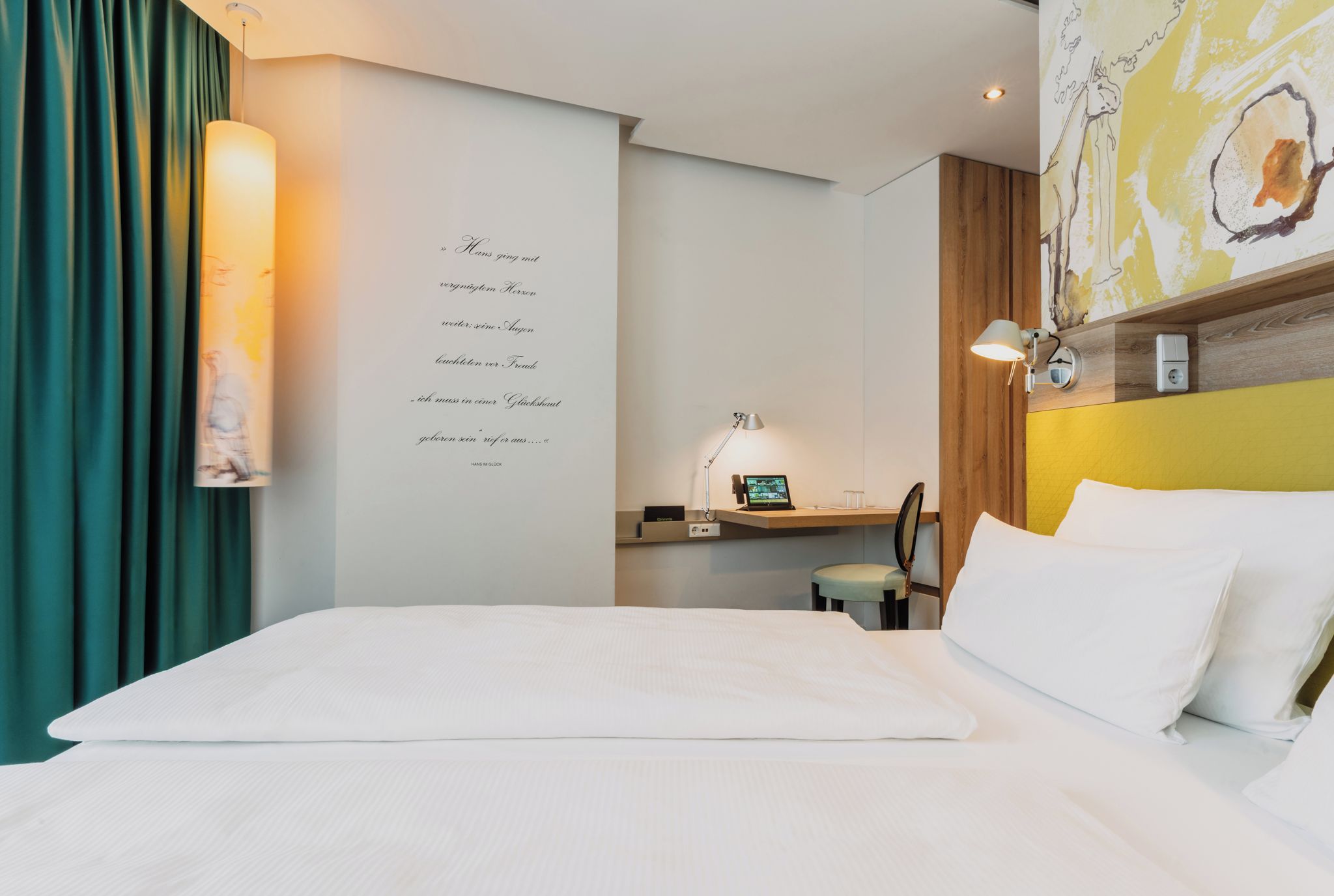 A hotel room with a bed, a desk with a computer, a lamp, and a wall with a decorative piece.