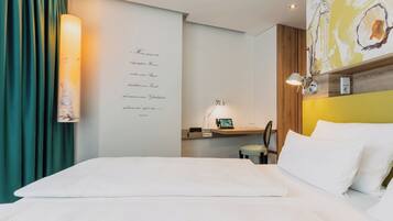 A hotel room with a bed, a desk with a computer, a lamp, and a wall with a decorative piece.