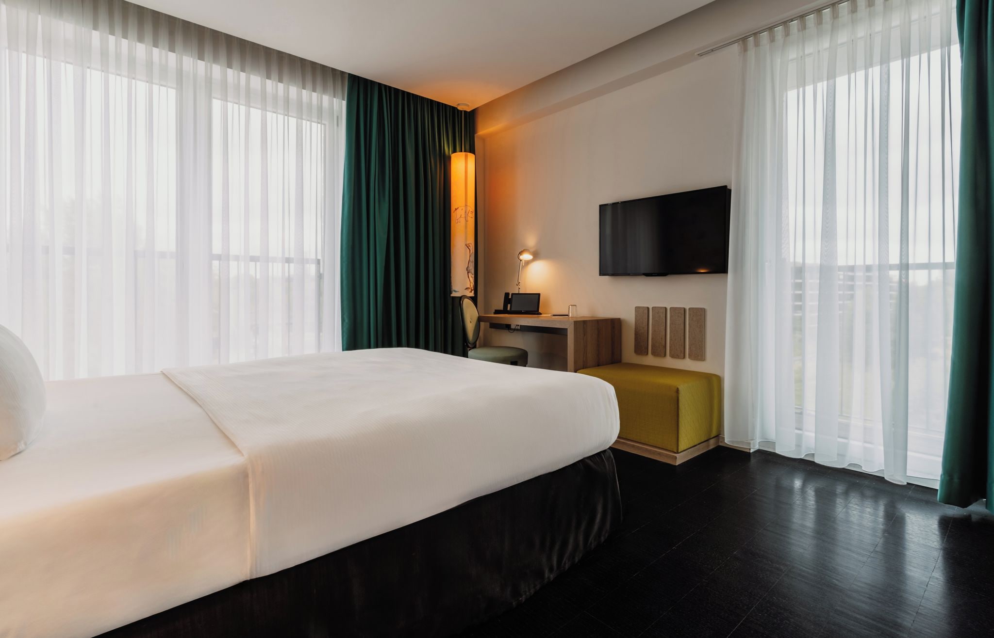 A hotel room with a bed, a desk with a laptop, a TV mounted on the wall, and a window with sheer curtains.