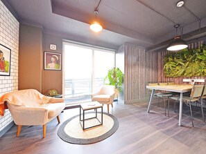 A modern living room with a sofa, armchair, coffee table, and dining table with chairs. There is a large window providing natural light and a view of the outdoors. -  For Students Only Ensuites in Birmingham (Birmingham)