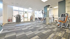 Une salle de sport équipée de divers appareils d’exercice, dotée d’un tapis à motifs et de grandes fenêtres laissant entrer la lumière naturelle.