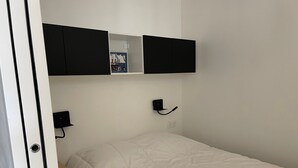 Una habitación pequeña y ordenada, con una cama, un estante en la pared con un cartel, y un enchufe.