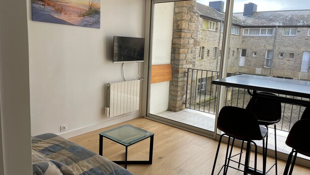 Una sala moderna con un sofá, una mesa de centro de vidrio, un televisor montado en la pared, un radiador y un balcón con mesa y sillas.