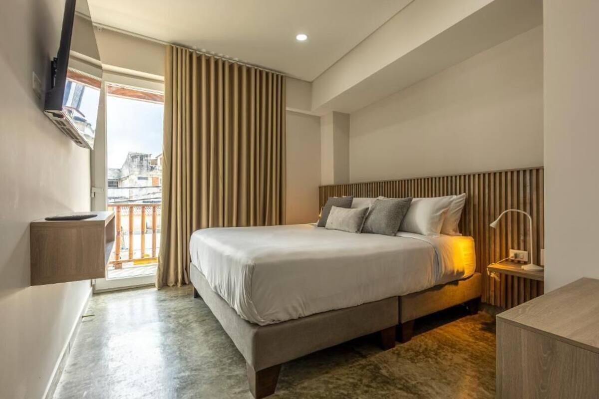 Una habitación de hotel moderna con una cama grande, una mesita de noche, un televisor montado en la pared y vistas a la ciudad a través de la ventana.