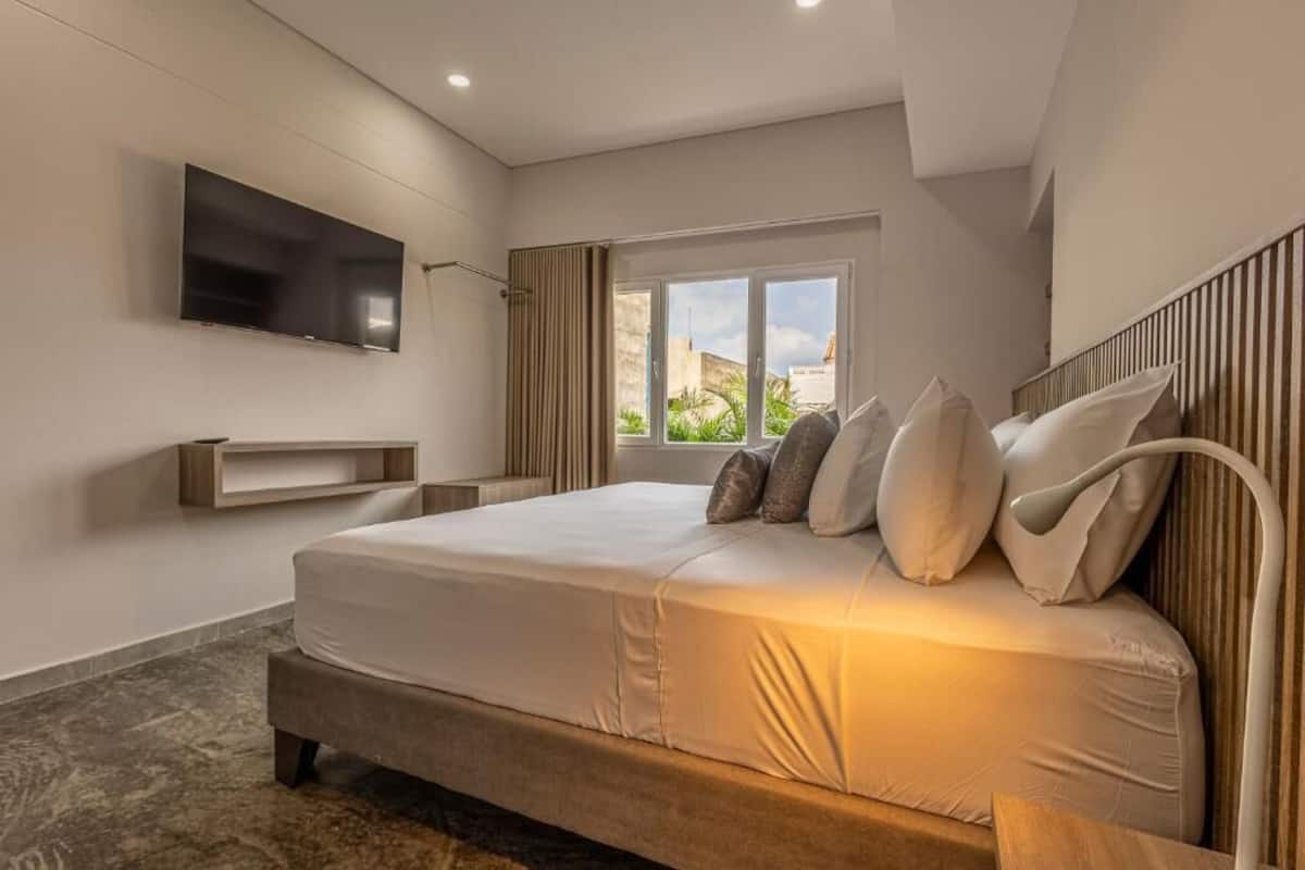 Una habitación de hotel moderna con una cama grande, un televisor de pantalla plana montado en la pared, una ventana con vistas al exterior y un banco empotrado debajo de la cama.