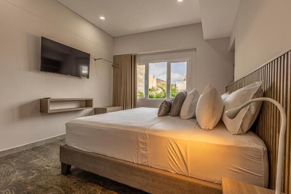 A modern hotel room with a large bed, a flat-screen TV mounted on the wall, a window with a view of the outside, and a built-in bench under the bed. - De la Cruz Studio apartments (Cartagena)