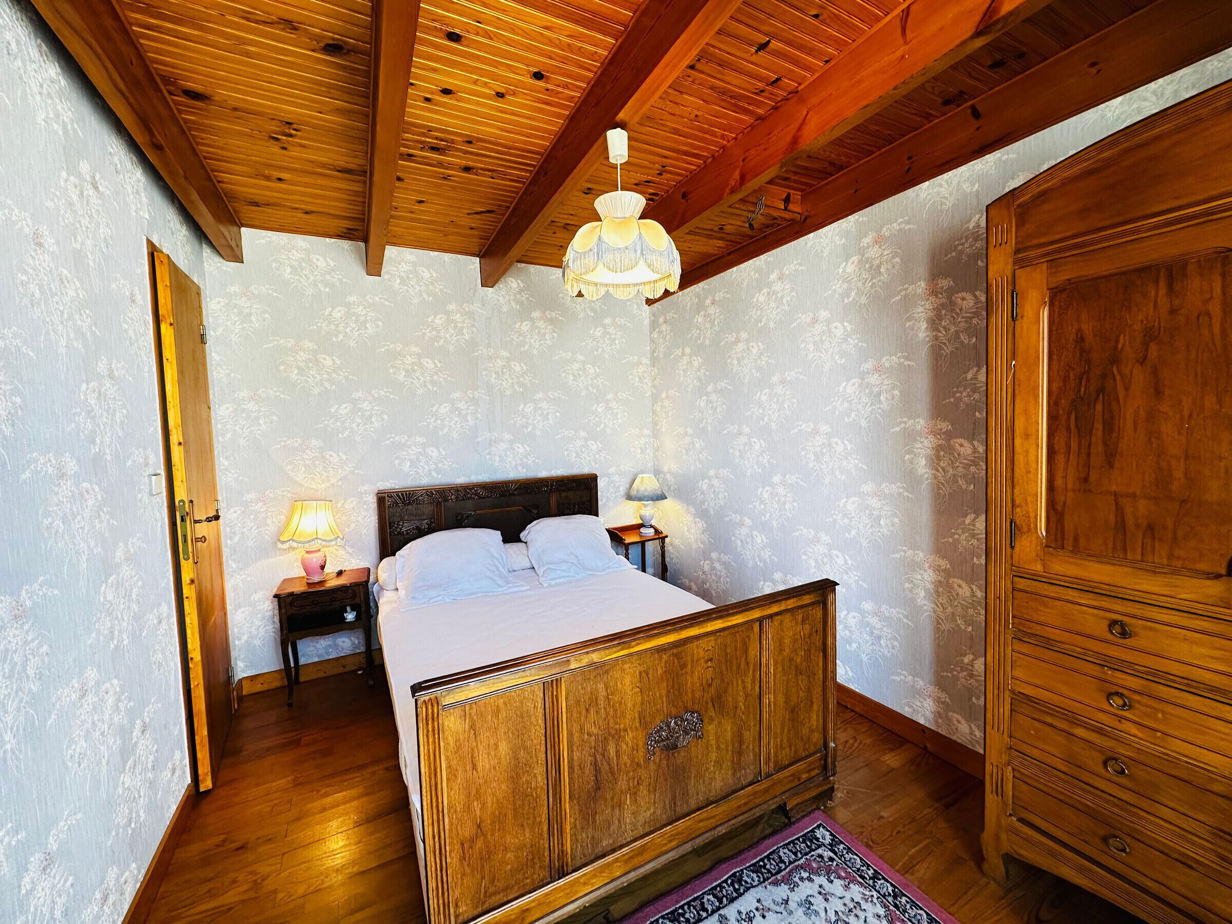 A bedroom with a wooden bed, a nightstand with lamps, a wardrobe, and a patterned wallpaper.