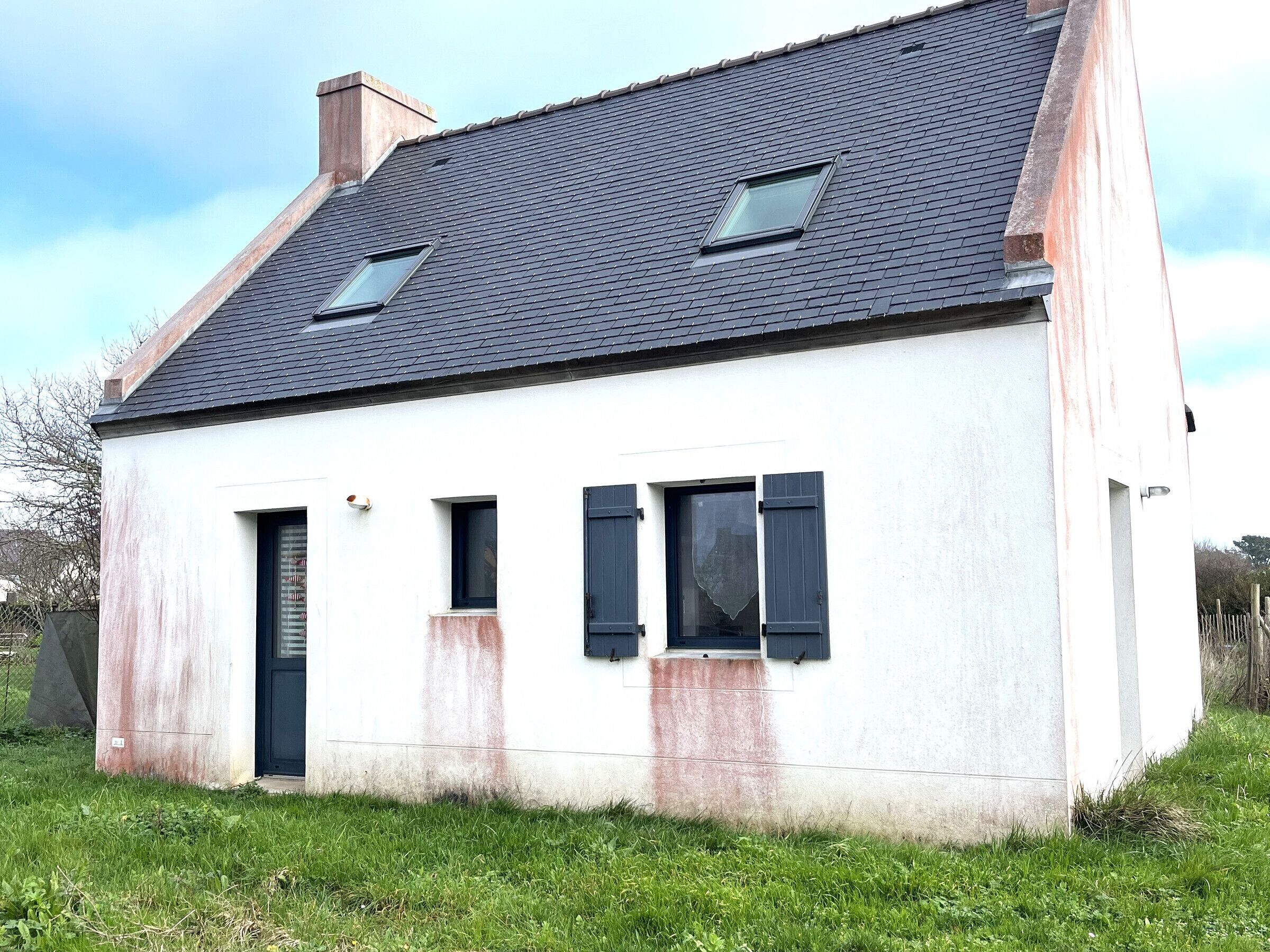 A single-story house with a sloped roof, two windows with shutters, and a visible chimney.
