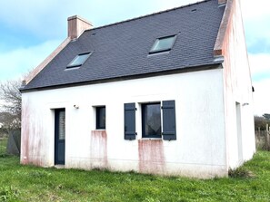 Una casa de un piso con techo inclinado, dos ventanas con persianas y una chimenea visible.