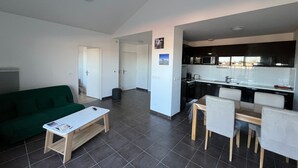 Ein modernes Apartment mit einem grünen Sofa, einem Holztisch, einer Essecke mit Stühlen, einer Küche mit schwarzen Schränken und einem Fliesenboden.