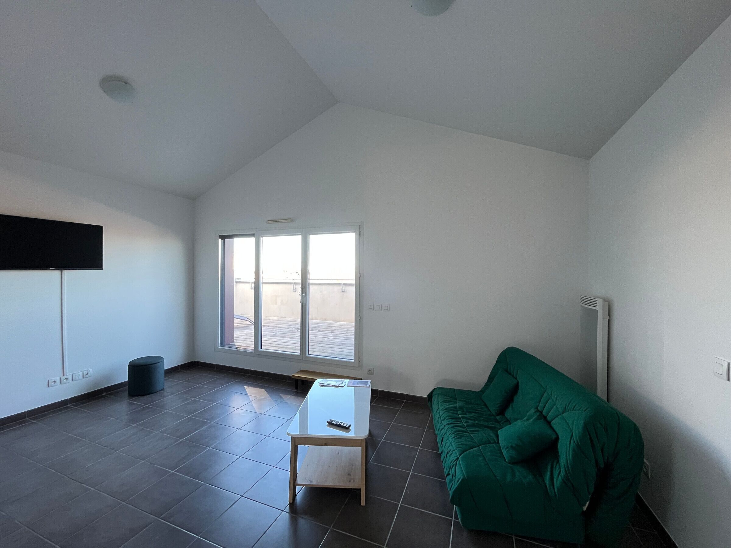A room with a large window, a green sofa, a small table, and a flat-screen TV mounted on the wall.