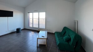 Ein Zimmer mit einem großen Fenster, einem grünen Sofa, einem kleinen Tisch und einem an der Wand montierten Flachbildfernseher.