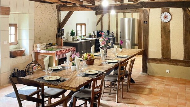A dining area with a long wooden table set for six, surrounded by chairs. A kitchen is visible in the background with modern appliances. - Charming country house in Loire - pers. (Monthodon)