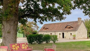 A rustic stone building with a tiled roof, a large tree, and outdoor seating with red cushions. - Charming country house in Loire - pers. (Monthodon)
