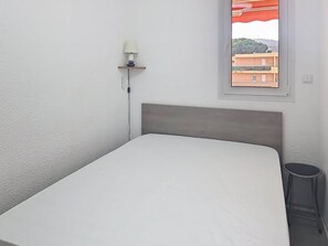 Ein kleines Zimmer mit einem Einzelbett, grauem Kopfteil, einer Nachttischlampe und einem Fenster mit Blick auf Gebäude.