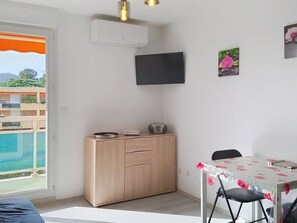 Ein Zimmer mit einem Holzkasten, einem Tisch mit floralem Tischläufer, einem an der Wand befestigten Fernseher und einem Balkon mit Blick auf Gebäude und Grünflächen.