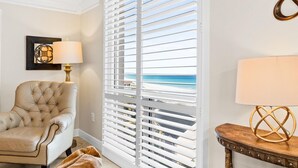 Une chambre avec vue sur la plage, un fauteuil à capitons, une table avec une lampe et une grande fenêtre avec des stores.
