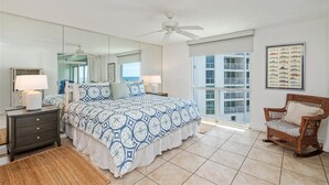 A bedroom with a large bed, a ceiling fan, a nightstand, a chair, a window with a view of a building, and a framed fish chart on the wall.