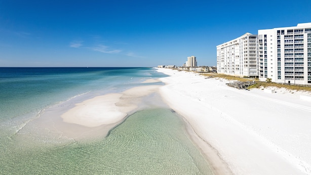 Ein Strand mit klarem, flachem Wasser, einem Sandufer und einer Reihe weißer Gebäude mit mehreren Etagen und Balkonen.