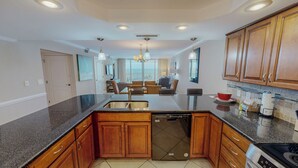 A modern kitchen with granite countertops, wooden cabinets, a dishwasher, and a dining area with wicker chairs. - Great Rate and Gulf View in ST 3064 (Destin)
