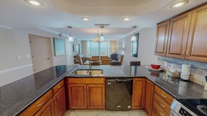 A modern kitchen with granite countertops, wooden cabinets, a dishwasher, and a dining area with wicker chairs. - Great Rate and Gulf View in ST 3064 (Destin)