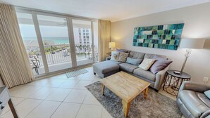 A living room with a sofa, a coffee table, a lamp, and a view of the ocean through a sliding door. - Great Rate and Gulf View in ST 3064 (Destin)