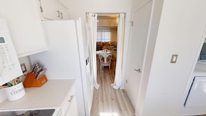 Une cuisine étroite avec des meubles blancs, un four à micro-ondes et un porte-ustensiles. Une porte donne sur un espace de vie comprenant un canapé et des coussins à motifs.