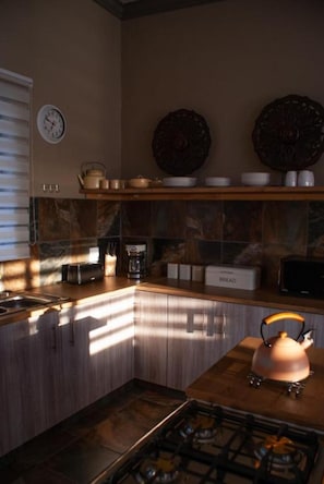 Una cocina con estufa a gas, tetera y panera. Hay un reloj en la pared y una repisa con platos y tazones.
