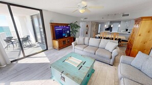 A spacious living area with a large sliding glass door leading to a balcony, a television, a sofa, a wooden cabinet, and a dining area with a table and chairs. - Gulf Front Views from Mainsail 264 (Miramar Beach)