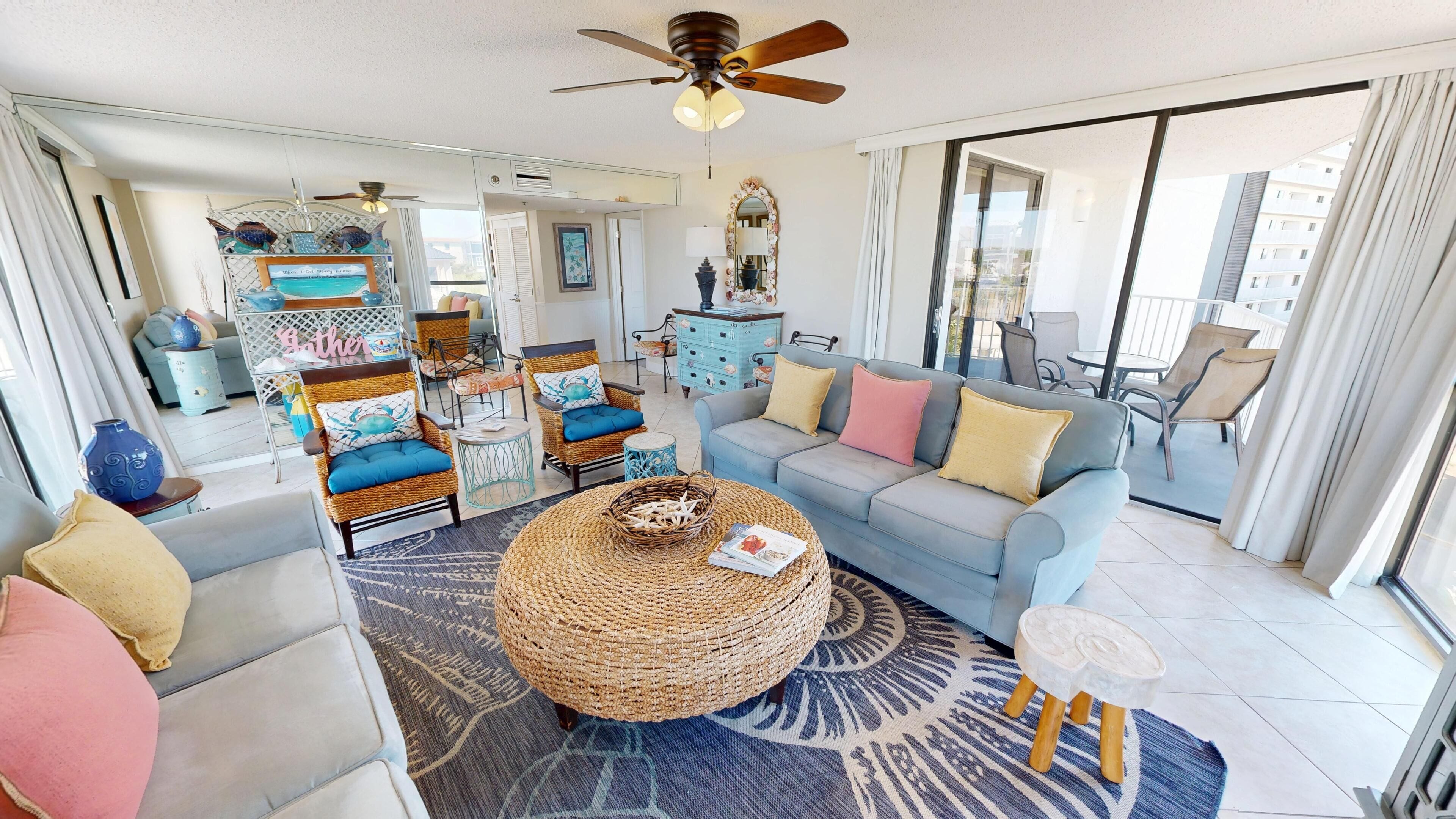 A spacious living room with a large grey sofa, wicker coffee table, and wicker chairs. There is a ceiling fan, a large mirror, and a view of the outside balcony.