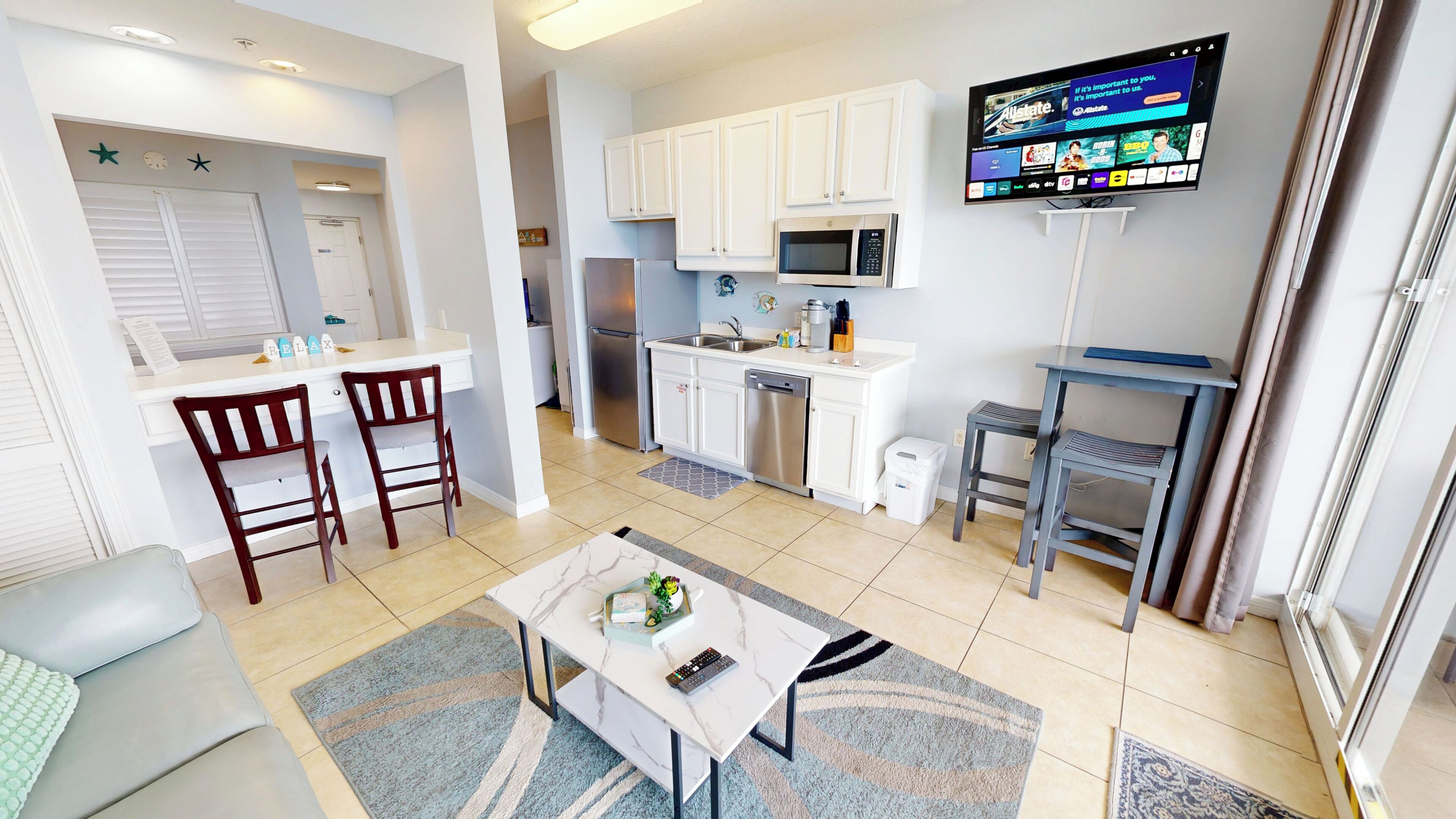 A modern kitchen with white cabinets, a stainless steel refrigerator, a microwave, and a flat-screen TV mounted on the wall.