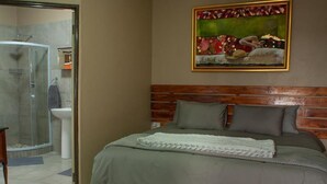 Une chambre avec une tête de lit en bois, un lit recouvert d’une couverture grise, un tableau accroché au mur et un espace douche.
