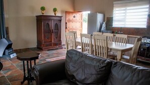 Een woonkamer met een zwarte leren bank, een houten eettafel en stoelen. Er staat een houten kast met twee potplanten erop. In de achtergrond zijn een koelkast en een fornuis zichtbaar.