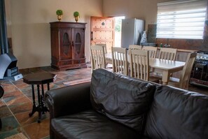 A living room with a black leather sofa, wooden dining table, and chairs. There is a wooden cabinet with two potted plants on top. A refrigerator and a stove are visible in the background.