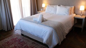Een net opgemaakt bed met witte lakens en een bedsprei met patroon, twee nachtlampjes en een rood vloerkleed met dessin.