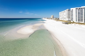 A beach with clear shallow waters, a sandy shore, and a row of white buildings with multiple floors and balconies. - Shoreline Towers 3103 Free Beach Setup (Destin)