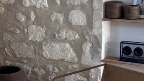 A stone wall with a rustic aesthetic, a woven chair, and a shelf with decorative items. - Petit Mas des Cigales Saint-Rémy de Provence (Saint-Rémy-de-Provence)