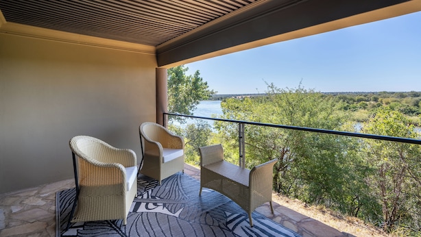 A balcony with wicker furniture and a patterned rug, overlooking a scenic landscape with trees and a body of water. - Simanya River Lodge (Nkurenkuru)