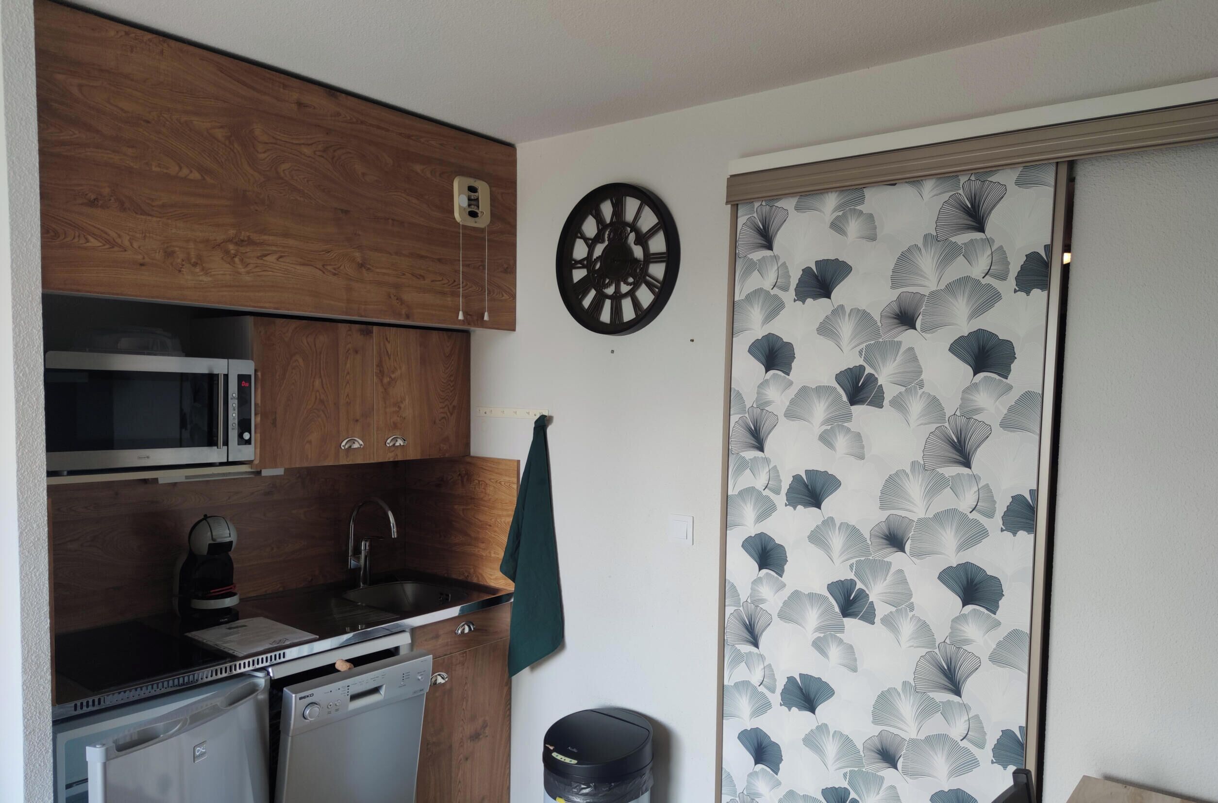Una cucina compatta con mobili in legno, un forno a microonde, un lavandino e una sveglia appesa al muro.