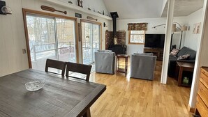 A spacious living area with a high ceiling, wooden flooring, a dining table with chairs, a fireplace, a television, and a kitchen area. - WATERFRONT ON SAND POND WITH A PRIVATE SANDY BEACH (East Baldwin)