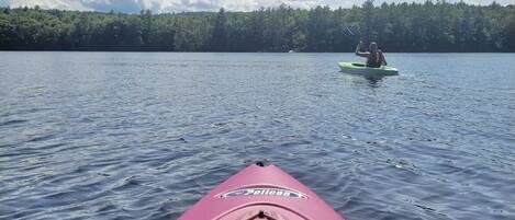 A pink kayak with a bottle and a whistle on the bow, heading towards a person in a green kayak on a calm lake with a forested shoreline.