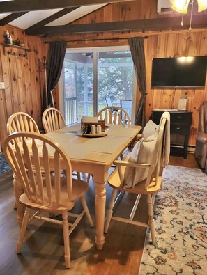 A wooden-paneled room with a dining table, chairs, a television, and a view of trees through large windows.