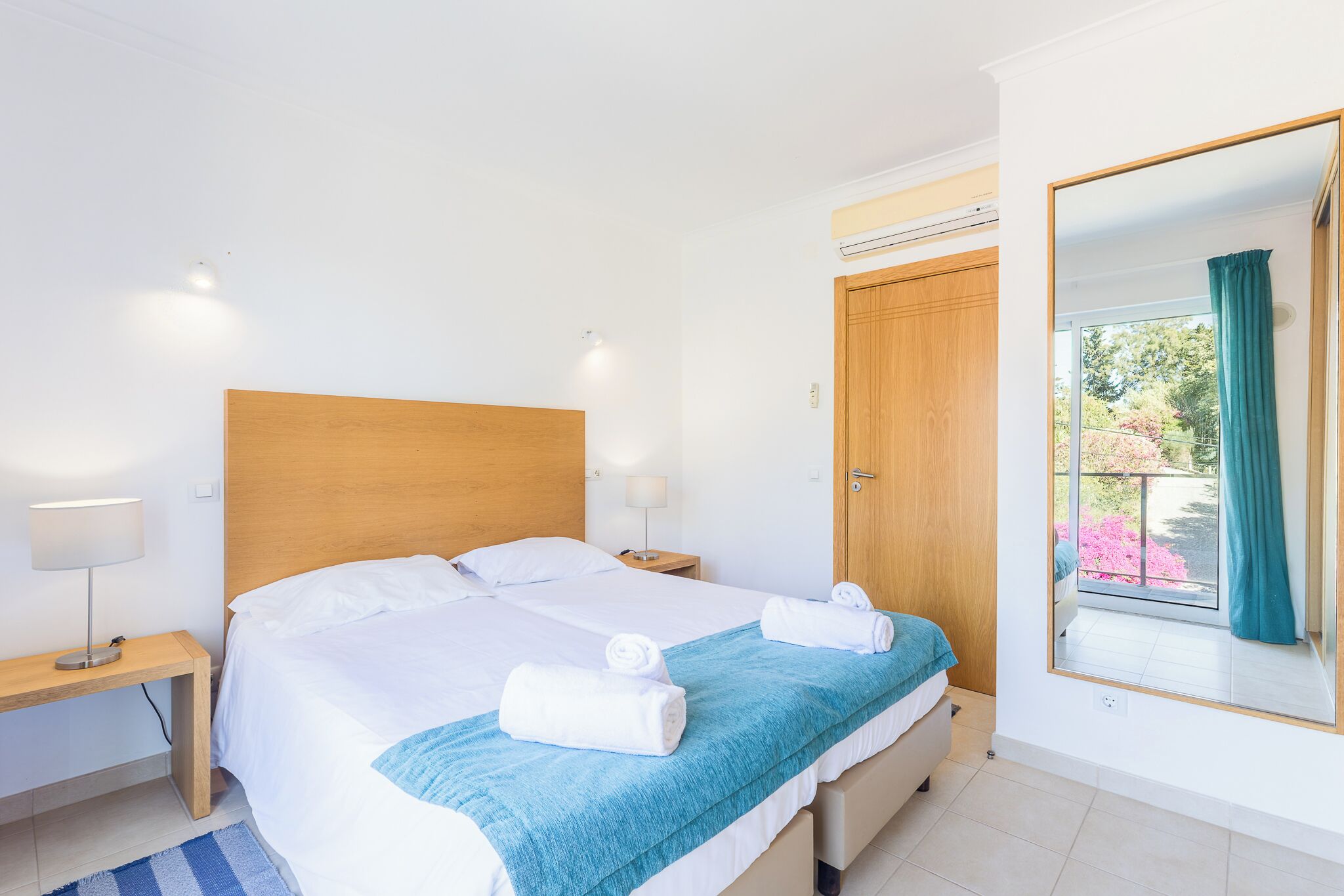 A hotel room with a bed, a nightstand, a mirror, and a window with a view of greenery.