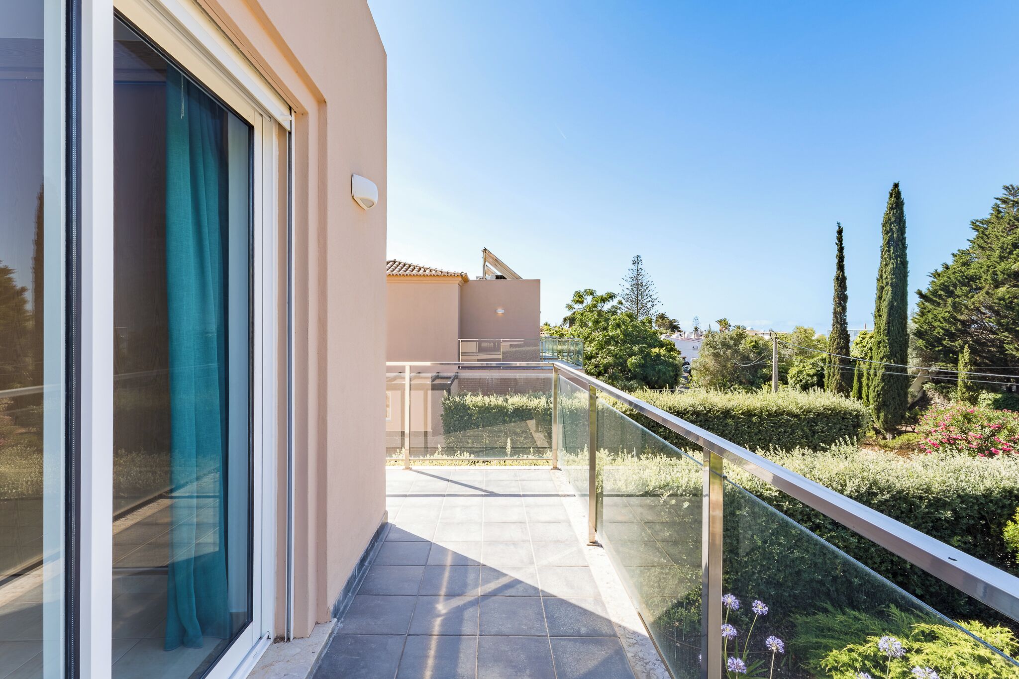 A balcony with a glass railing, a sliding glass door, and a view of a landscaped garden with trees and shrubs.